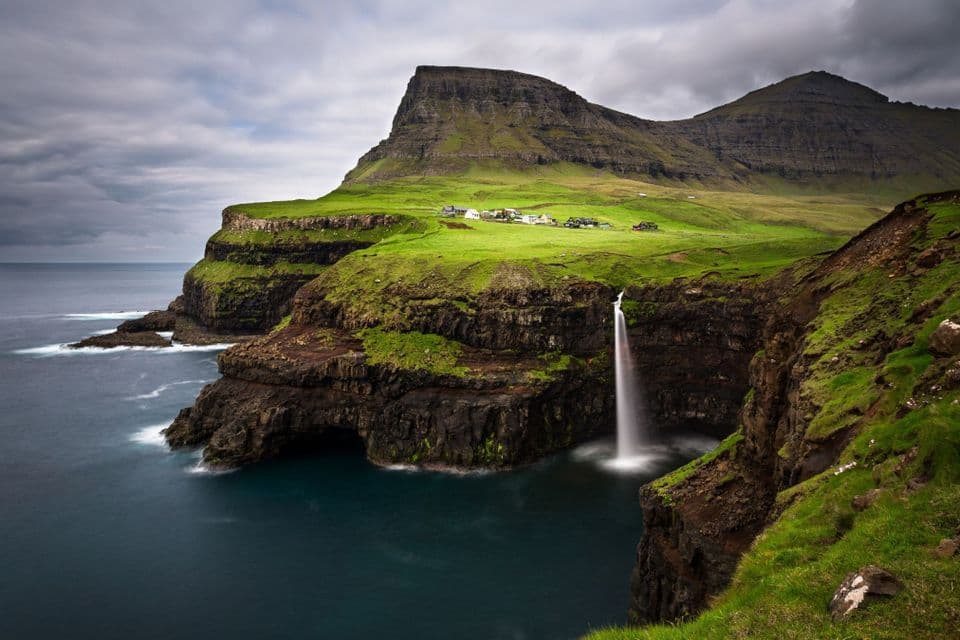 Una cascata scende da una verde scogliera erbosa nell'oceano, con un piccolo villaggio visibile in cima alla scogliera sotto un cielo nuvoloso.