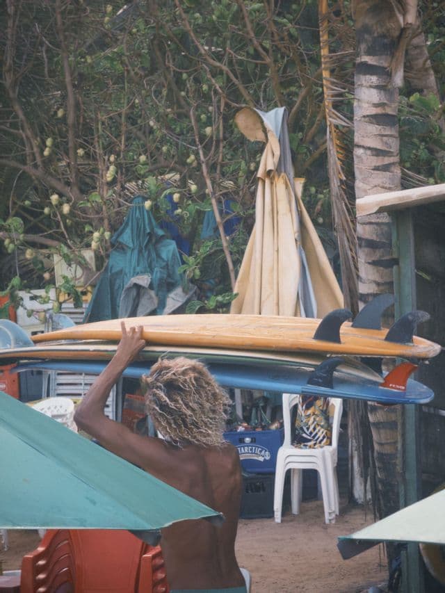 Una persona vista di spalle, con capelli biondi e ricci, bilancia due tavole da surf impilate sulla testa su una spiaggia sabbiosa con palme.