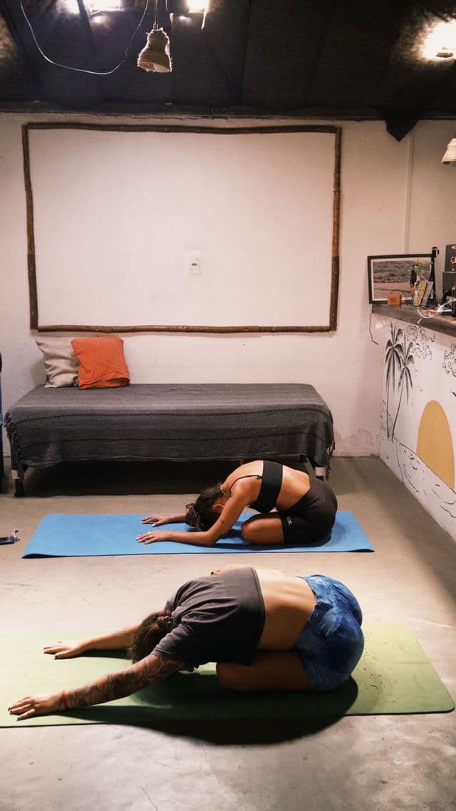 Due donne praticano yoga, tenendo la posizione del bambino su tappetini separati su un pavimento in cemento al chiuso.