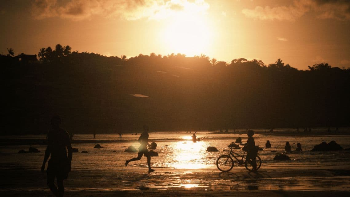 Sagome di persone su una spiaggia bagnata al tramonto, con una persona che corre e un'altra che cammina con una bicicletta.