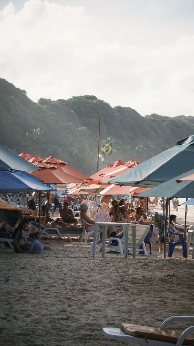 Persone si rilassano su una spiaggia di sabbia affollata sotto ombrelloni colorati, con bandiera brasiliana e collina verde sullo sfondo.
