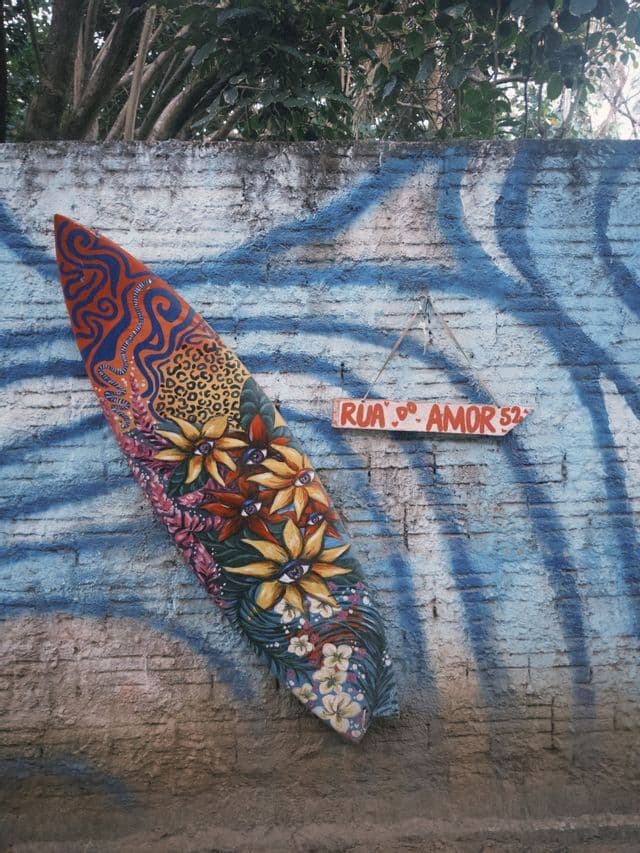 Una tavola da surf decorata con motivi colorati di fiori con occhi e stampe animali si appoggia a un muro di mattoni dipinto con onde blu.