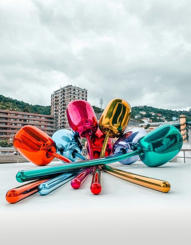 A colorful, metallic sculpture of tulips rests on a white platform against a city skyline under a cloudy sky.