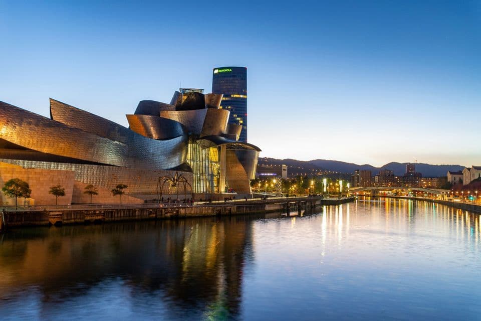 The illuminated exterior of a modern, metallic museum and a large spider sculpture reflected in a river at dusk.