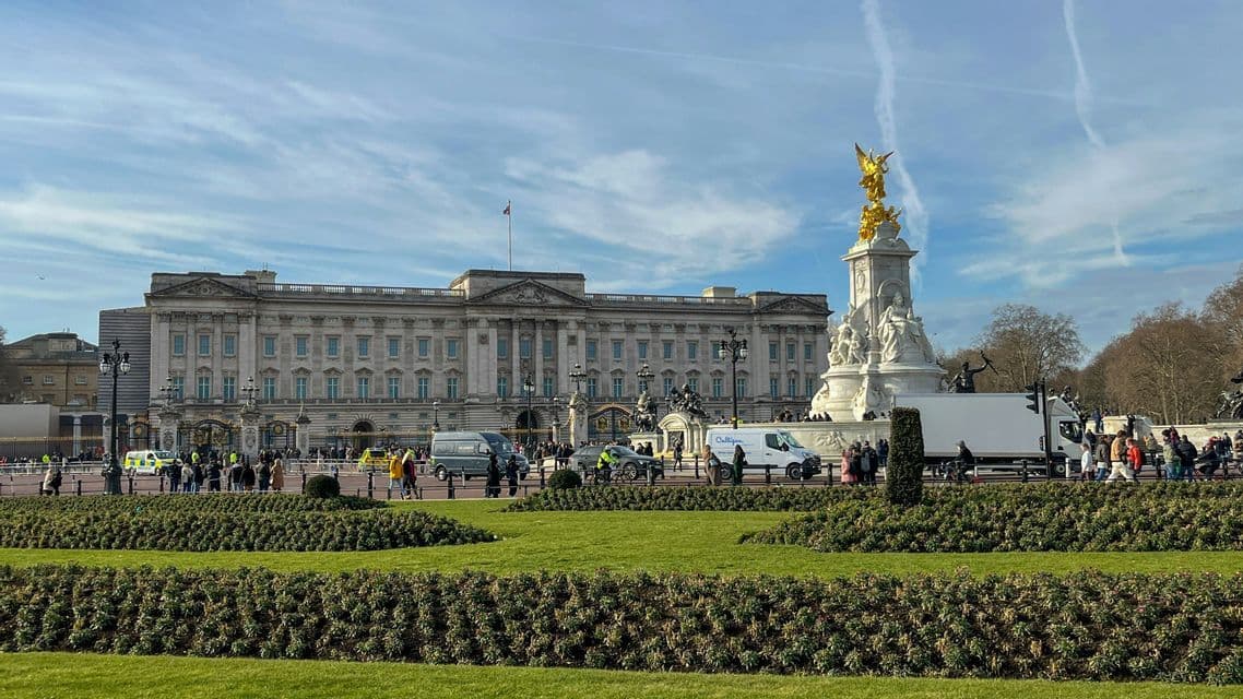 A grand classical palace and a golden-topped memorial statue stand behind a lawn and a road with people and traffic.