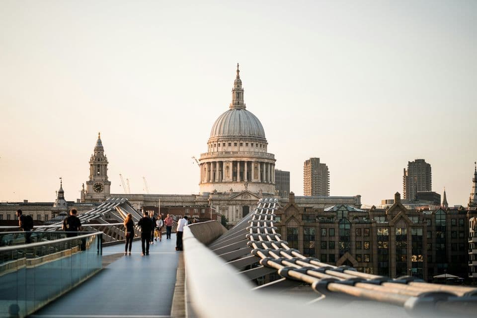 People walk on a modern pedestrian bridge towards a large, historic domed cathedral against a pale sky at sunset.