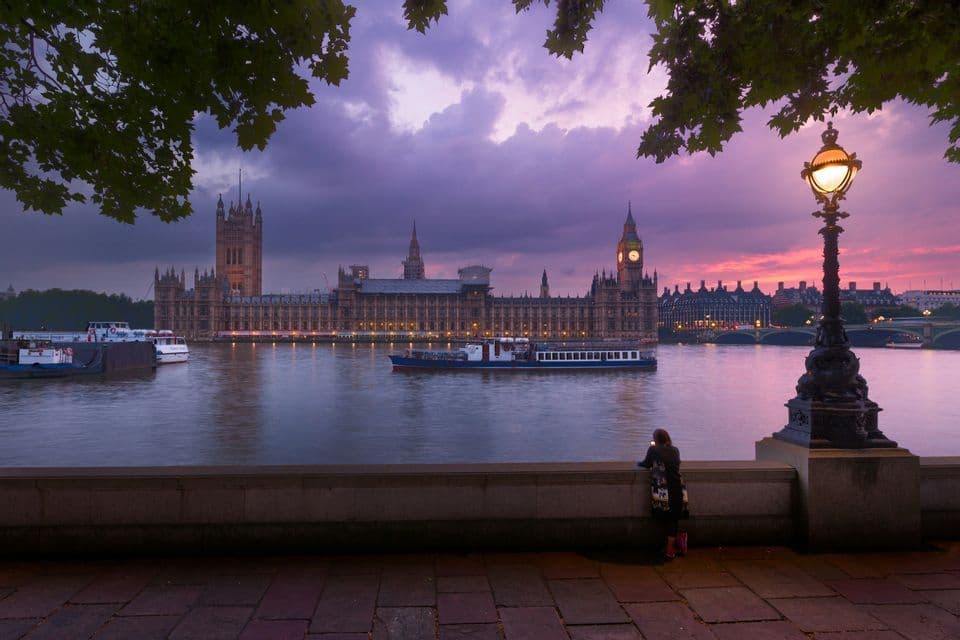 A person stands on a promenade looking at the Houses of Parliament across the River Thames, illuminated against a purple sunset sky.