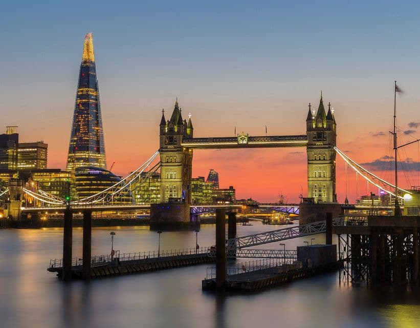 An illuminated historic bridge and a modern skyscraper standing over a river at sunset, with docks in the foreground.