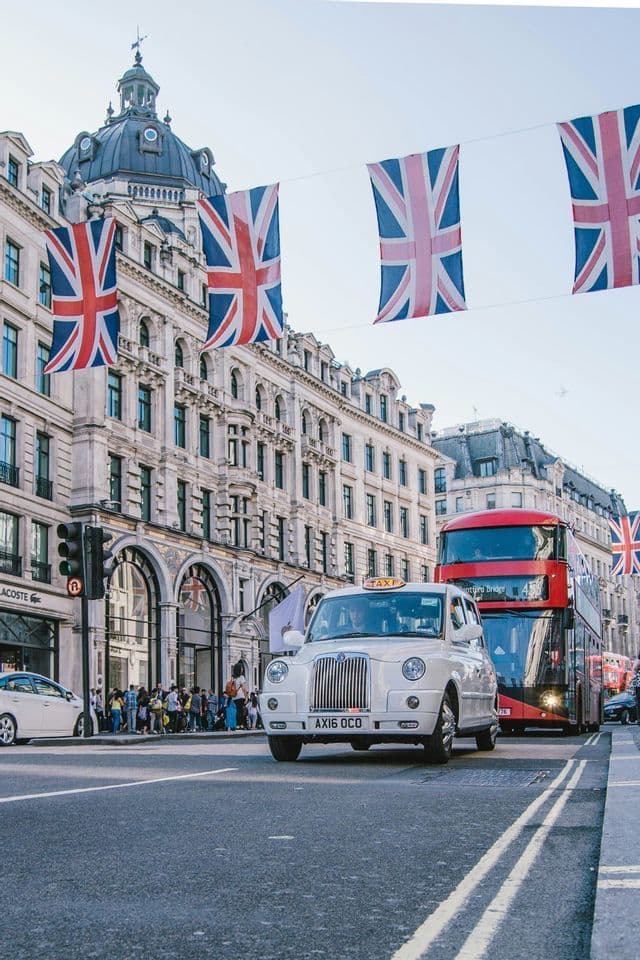 A white taxi and a red double-decker bus drive on a city street decorated with British flags hanging over the road.
