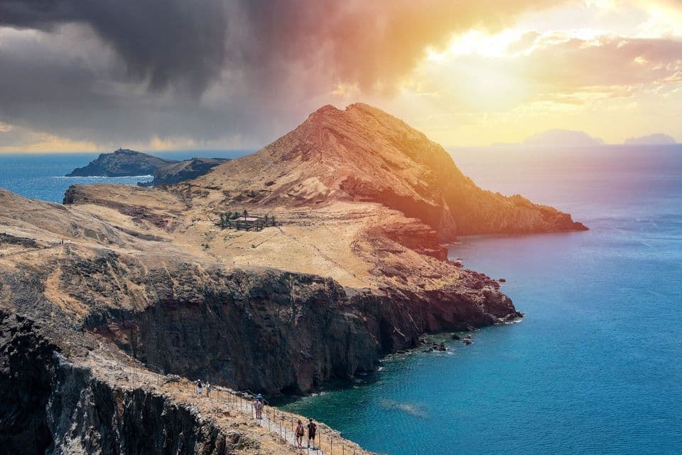 Un'escursione di gruppo WeRoad su un sentiero lungo una penisola scoscesa e rocciosa a picco sull'oceano, con la luce del sole che filtra tra le nuvole scure.