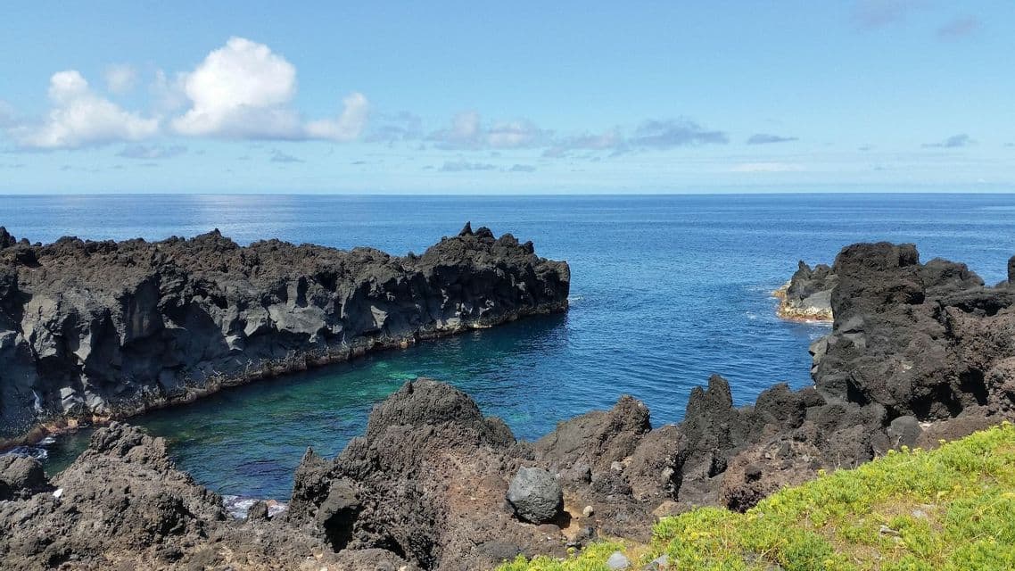 Scure rocce vulcaniche formano una piccola insenatura con acqua cristallina e blu su una costa sotto un cielo parzialmente nuvoloso.