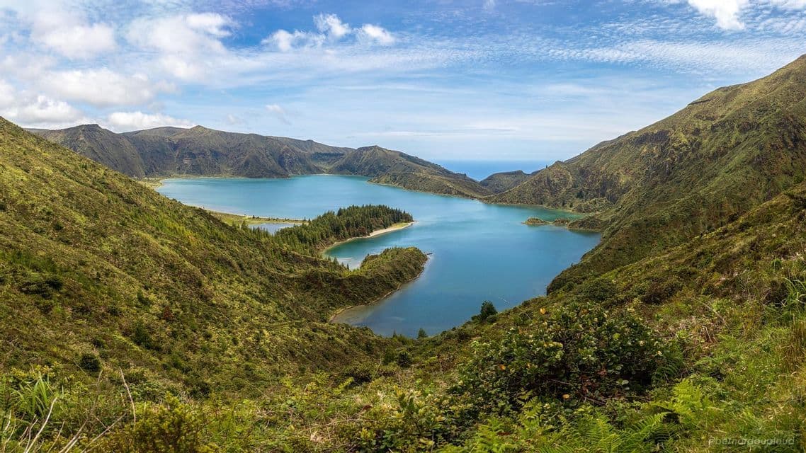 Un lago di cratere blu circondato da montagne verdi lussureggianti sotto un cielo parzialmente nuvoloso, con l'oceano visibile in lontananza.