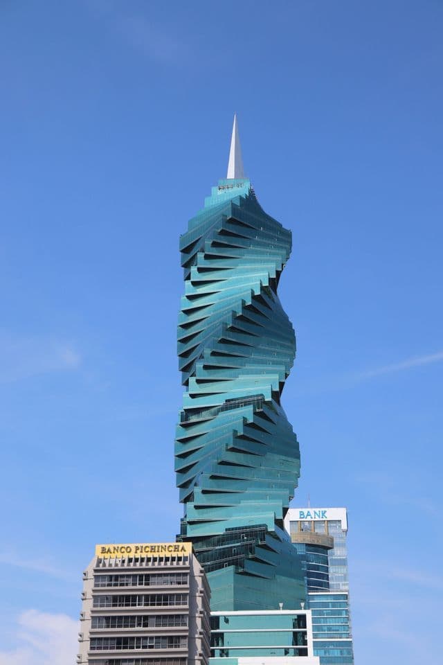 A modern skyscraper with a twisting teal glass facade and a spire rises against a clear blue sky above other city buildings.