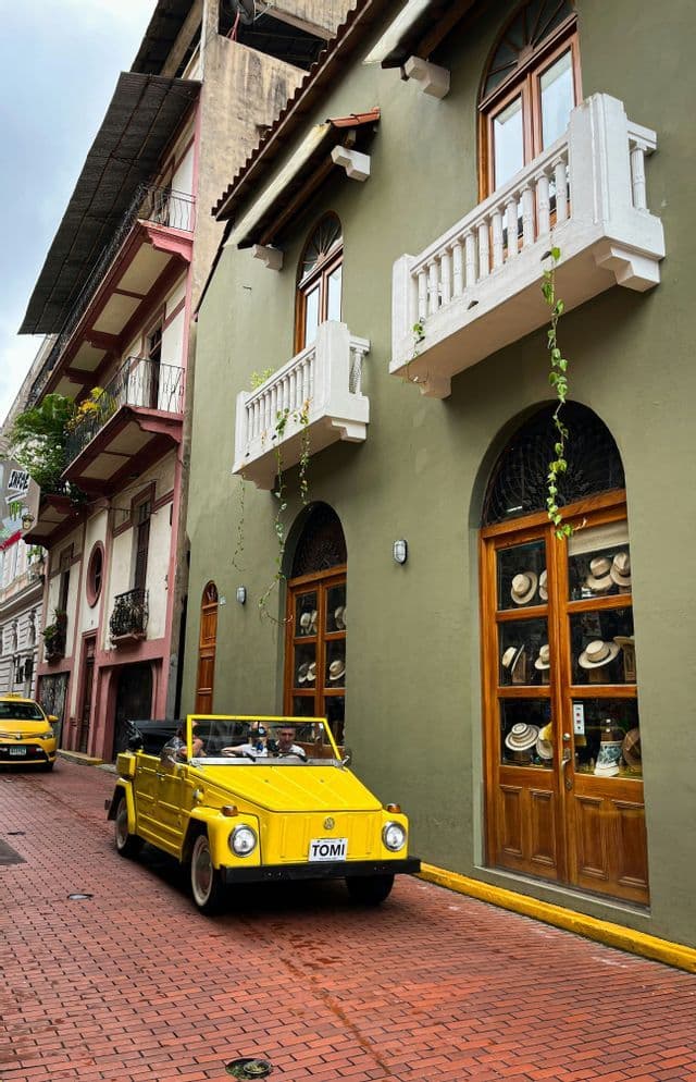 A yellow vintage convertible car drives down a narrow cobblestone street lined with colorful colonial buildings.