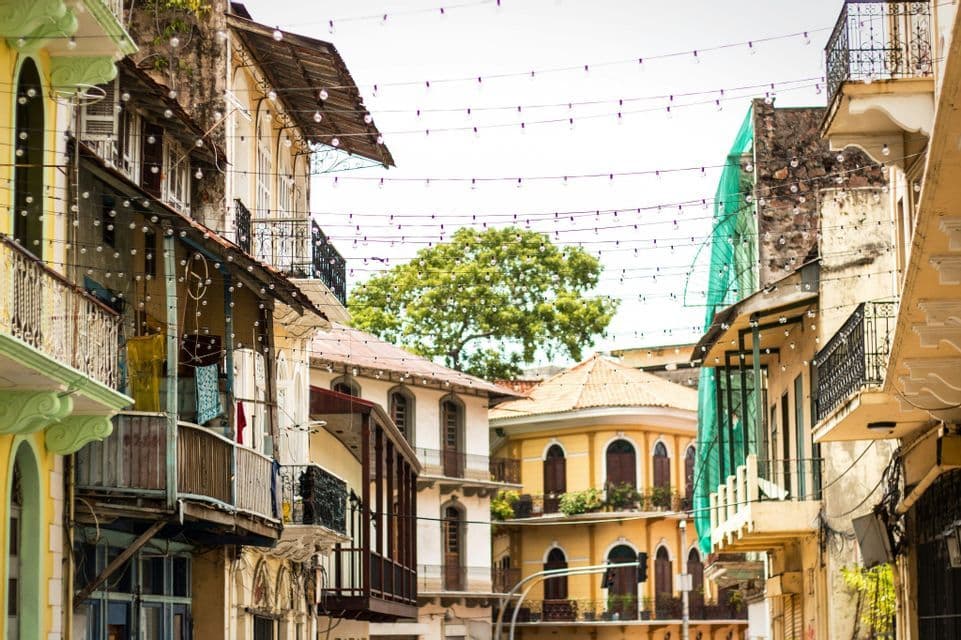 A view up a narrow street showing weathered colonial buildings with balconies, under strings of lights and a bright sky.