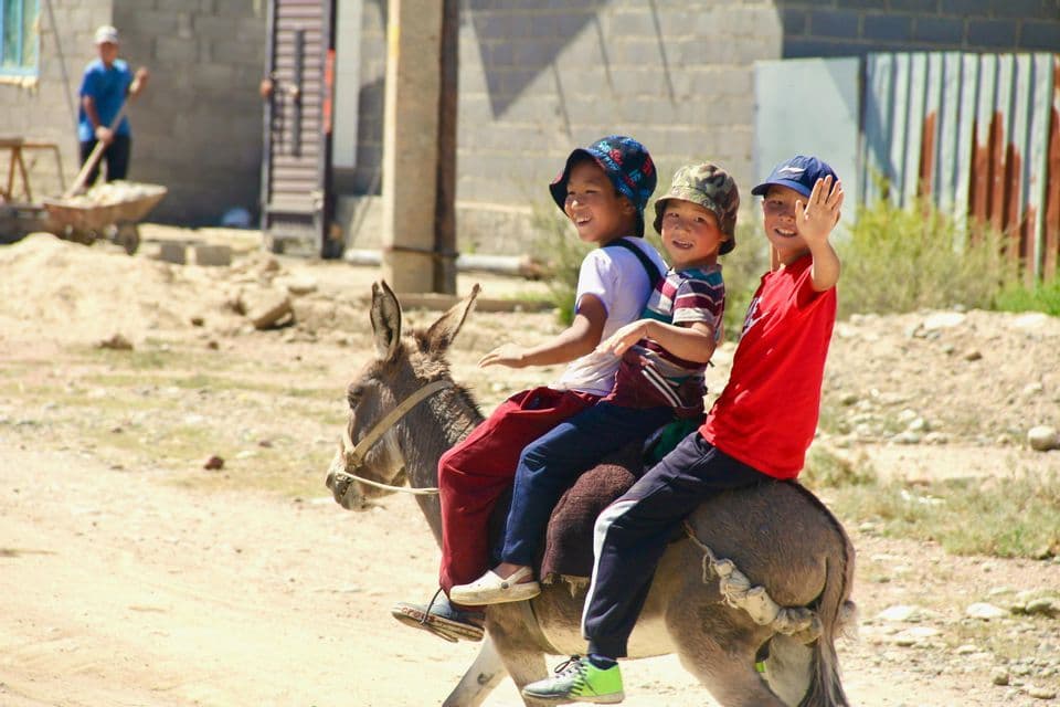 Trois garçons souriants chevauchent ensemble un âne sur un chemin de terre, l'un d'eux faisant signe à la caméra.