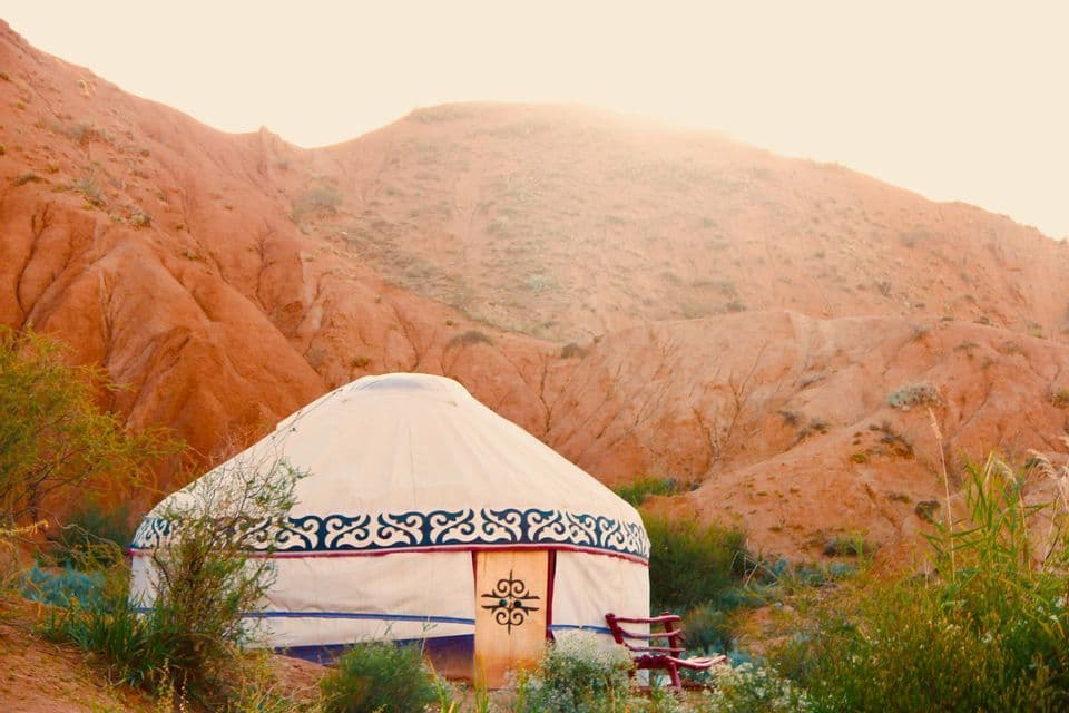 Une yourte blanche traditionnelle aux motifs décoratifs se dresse dans un paysage de collines de sable rouge et de végétation verte clairsemée.