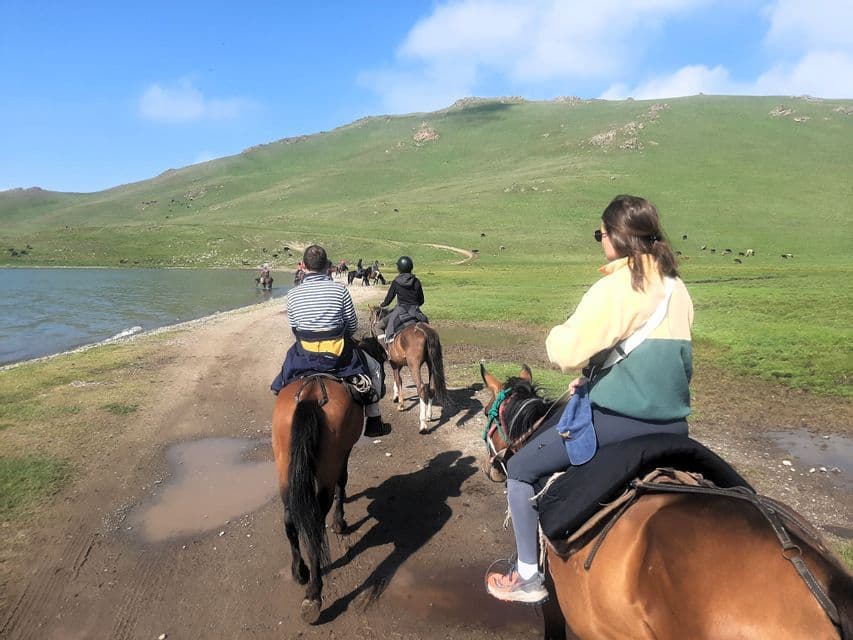 Un voyage de groupe WeRoad à cheval sur un chemin de terre à côté d'un lac, avec de grandes collines vertes en arrière-plan.