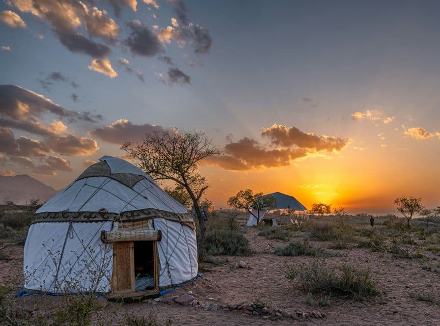 Une yourte blanche traditionnelle se dresse dans un paysage aride sous un coucher de soleil doré avec des nuages épars.