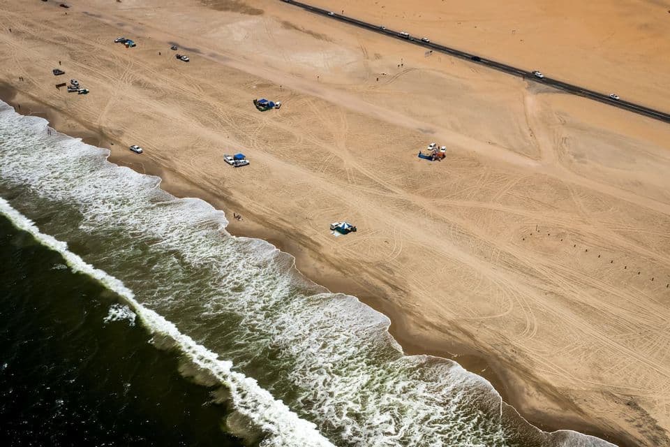 An aerial view of a sandy beach where the ocean meets the shore, with vehicles and tents on the sand and a road running parallel to the coast.