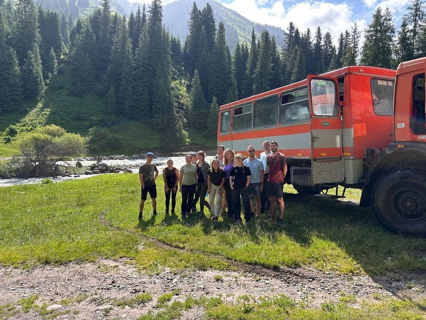 Un groupe WeRoad prend la pose devant un bus tout-terrain rouge dans une vallée de montagne verdoyante, traversée par une rivière.