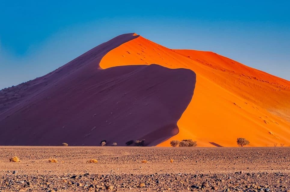 Una gran duna de arena naranja proyecta una sombra profunda sobre sí misma en un paisaje desértico rocoso bajo un cielo azul claro.