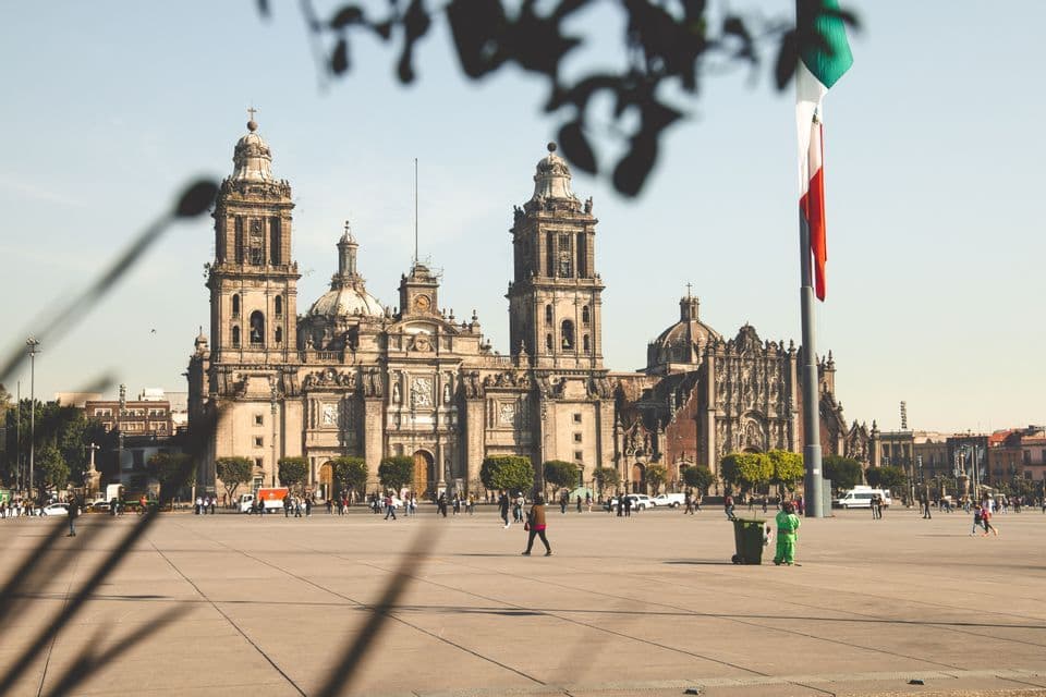 Una vista amplia de una catedral histórica y ornamentada, al otro lado de una plaza pública soleada, con una gran bandera mexicana ondeando en un poste a la derecha.