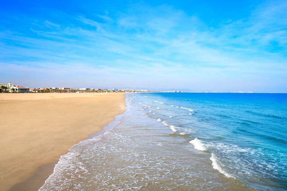 Une vaste plage de sable borde l'océan calme et bleu sous un ciel lumineux, avec un horizon urbain visible le long du littoral lointain.