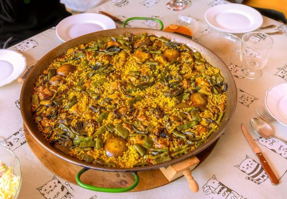 Une grande paella avec riz jaune, viande et légumes, posée sur une table dressée d'assiettes et de couverts.