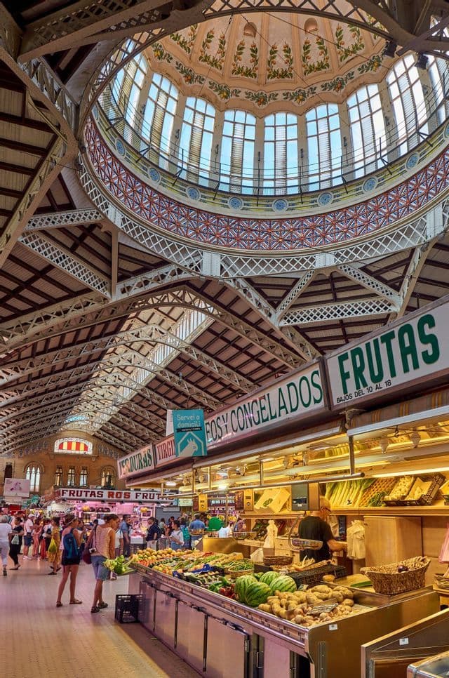 Un marché alimentaire couvert et animé, rempli de chalands sous un plafond en dôme orné de grandes fenêtres.