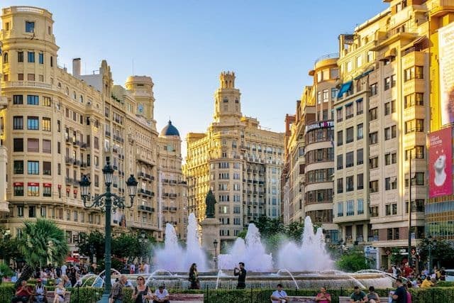 Une grande fontaine jaillit sur une place de ville animée, entourée de bâtiments historiques ornés, sous un ciel dégagé.