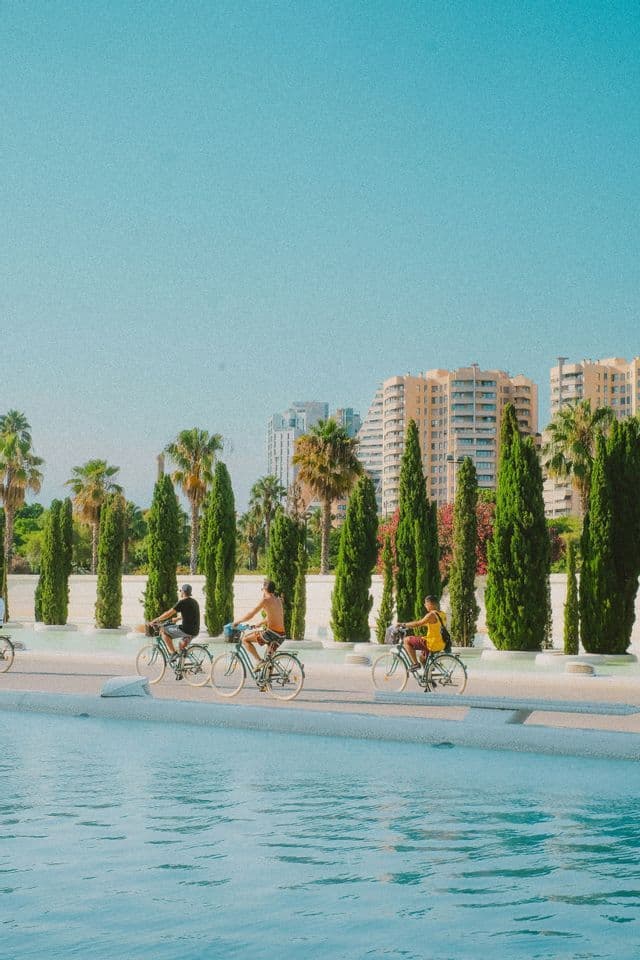 Un voyage de groupe WeRoad de trois hommes à vélo sur un chemin à côté d'une piscine, avec de grands arbres et des bâtiments urbains en arrière-plan.