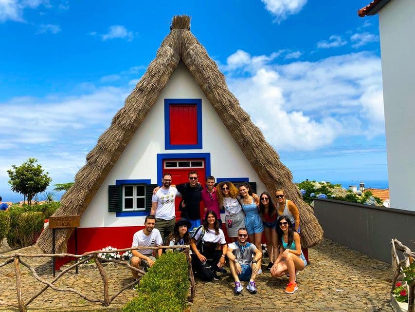 Un groupe WeRoad pose pour une photo devant une maison traditionnelle au toit de chaume en forme de A, par une journée ensoleillée.
