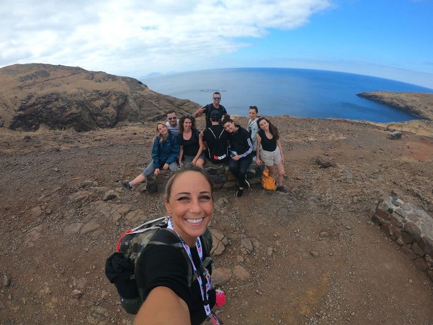A smiling woman takes a selfie with her WeRoad group trip on a rocky cliffside overlooking the sea under a cloudy blue sky.