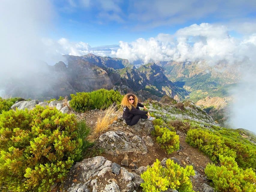 Una donna con i capelli ricci siede su una cima rocciosa di montagna, circondata da arbusti verdi, con vista su una vasta valle montana piena di nuvole.