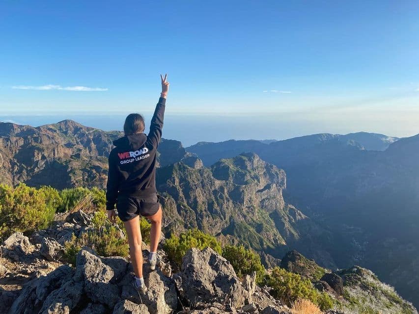 Un líder de grupo de WeRoad, de espaldas a la cámara, de pie en la cima de una montaña rocosa, haciendo el signo de la paz sobre una vasta cadena montañosa.