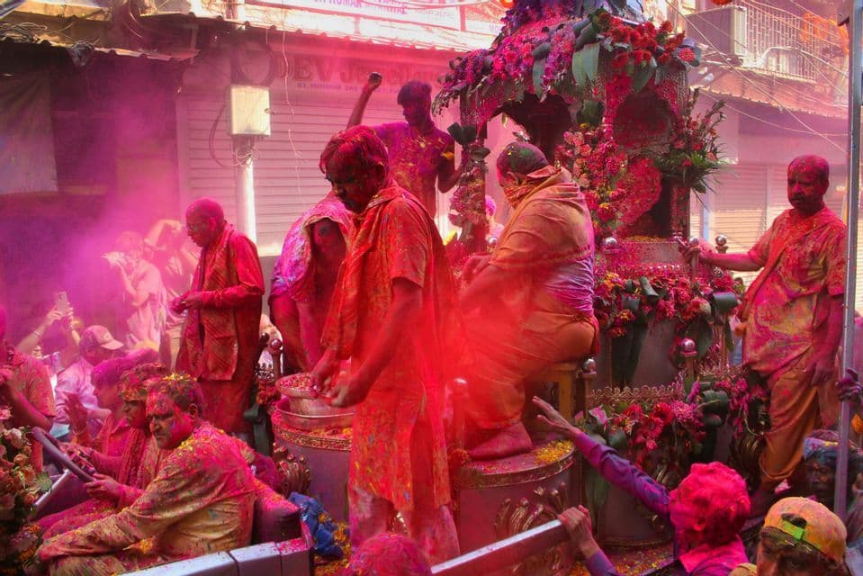 Una folla di persone festeggia su un carro allegorico decorato durante una festa di strada, avvolta in nuvole di polvere rossa e rosa.