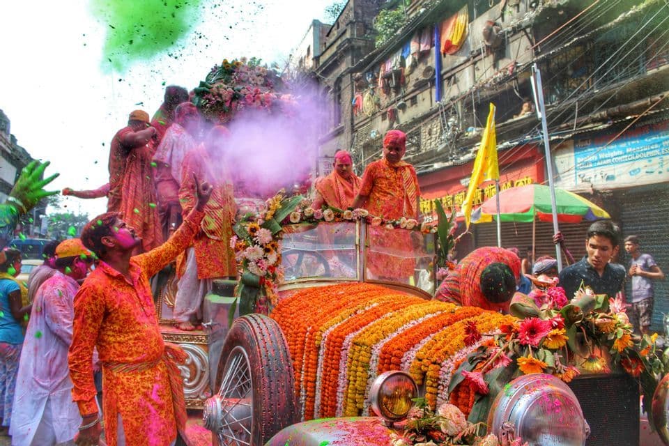 Un gruppo di persone ricoperte di polvere colorata festeggia su un'auto d'epoca decorata con ghirlande di fiori durante una parata di strada.