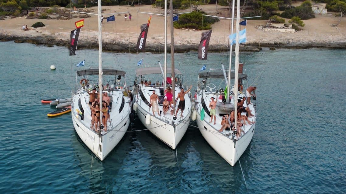 Eine WeRoad-Gruppe posiert auf drei weißen Segelbooten, die nebeneinander in einer ruhigen Bucht nahe einem bewaldeten Ufer ankern.