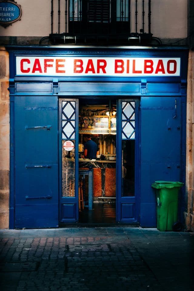 La facciata blu del Cafe Bar Bilbao su una strada acciottolata, con un'insegna illuminata e una porta aperta.
