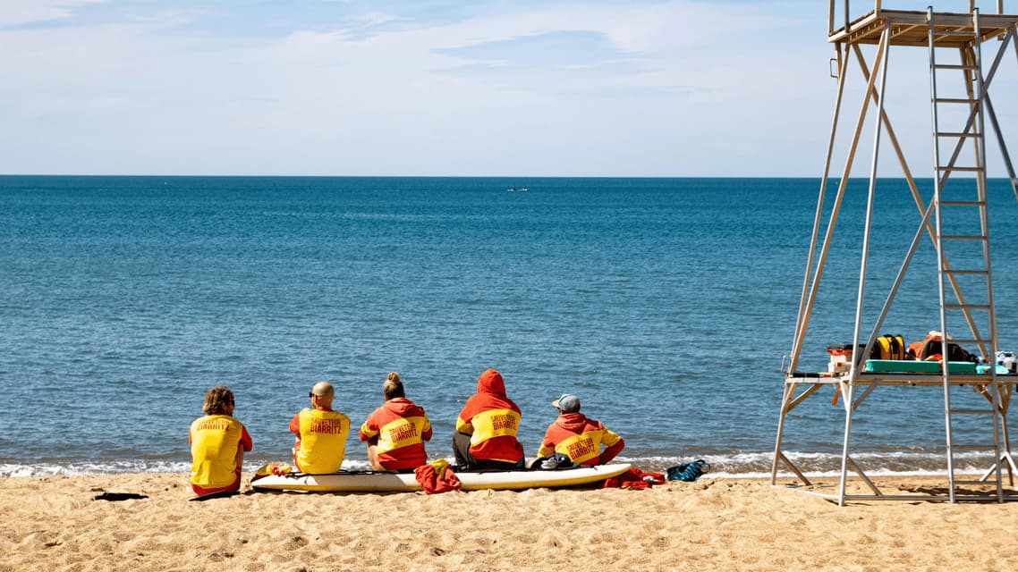 Cinque bagnini in uniformi rosse e gialle siedono su una tavola di salvataggio su una spiaggia sabbiosa, osservando l'oceano accanto a una torre di salvataggio.