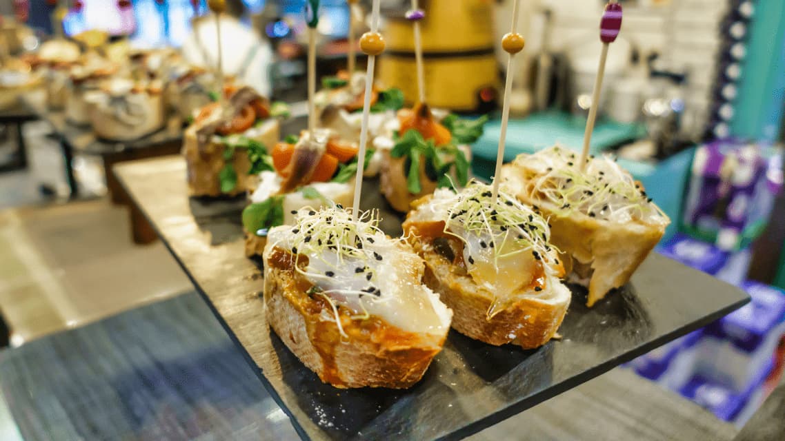 Un primo piano di pintxos spagnoli su un vassoio di ardesia nera, guarniti con germogli e semi neri, ciascuno fissato da uno spiedino.