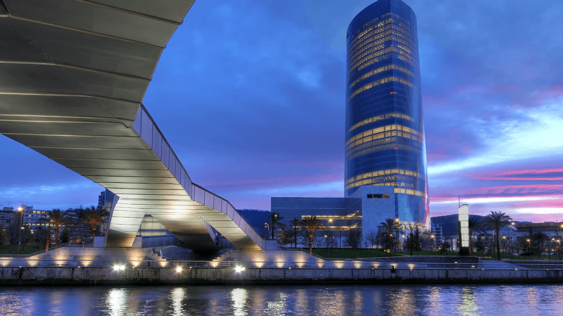 Un ponte moderno illuminato e un alto grattacielo cilindrico accanto a un fiume sotto un cielo crepuscolare.