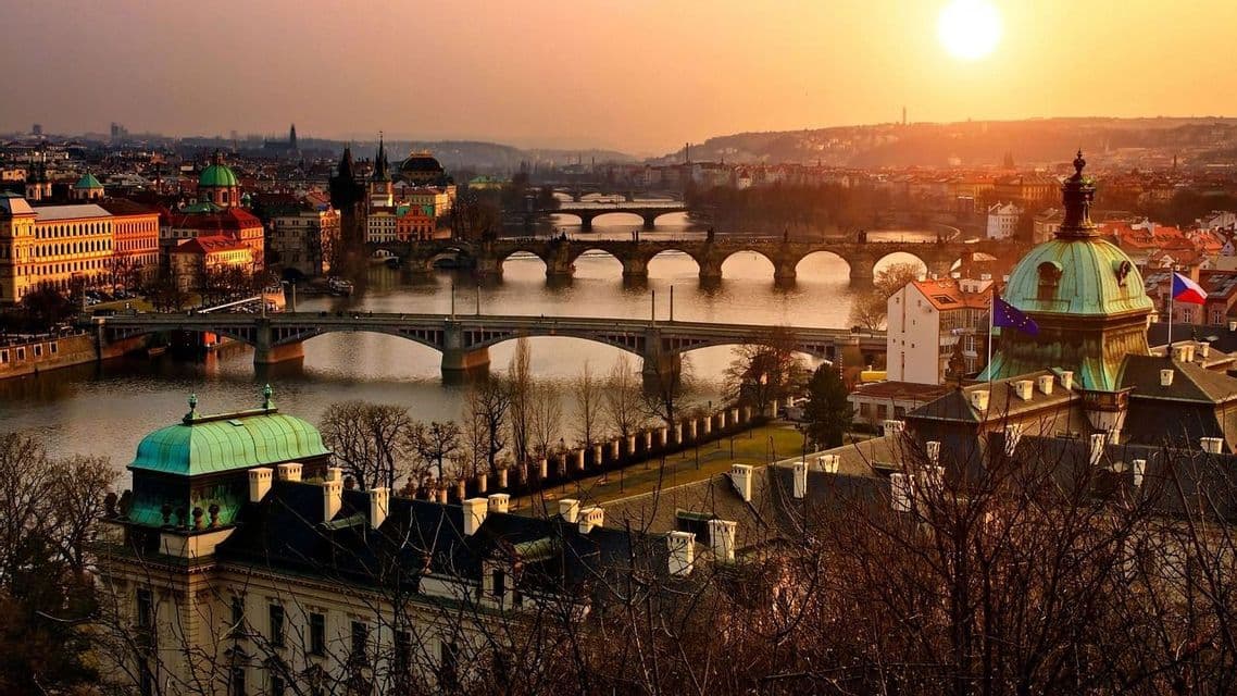 Vue plongeante sur un paysage urbain historique avec de nombreux ponts enjambant une rivière, baigné par la lumière chaude du soleil couchant.