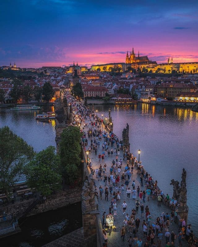 Vue aérienne d'un pont pavé bondé sur une rivière au crépuscule, avec un château illuminé sur une colline à l'arrière-plan.