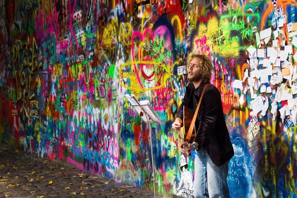 Un musicien de rue aux cheveux bouclés joue de la guitare acoustique et chante devant un long mur couvert de graffitis colorés.
