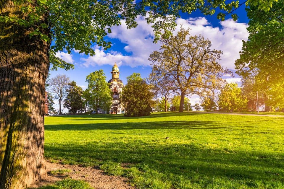 Un bâtiment historique avec une tour se dresse dans un parc verdoyant entouré d'arbres sous un ciel lumineux et nuageux.