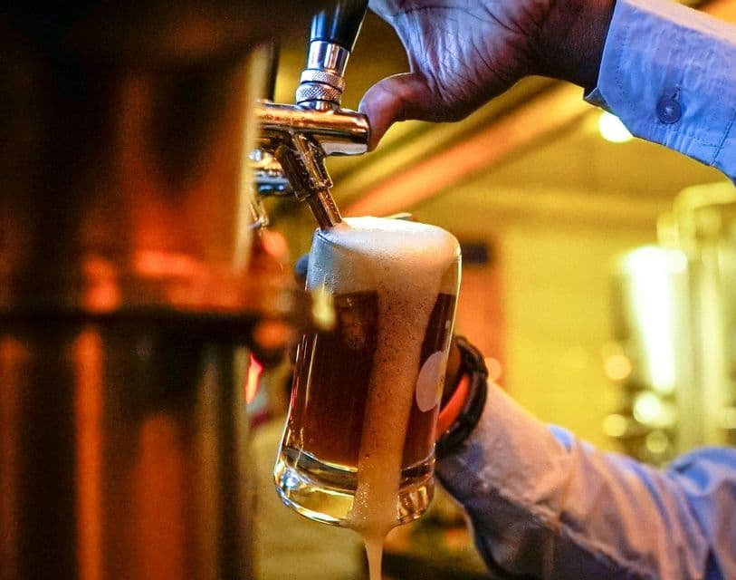 Gros plan sur une main versant une bière brune depuis un robinet métallique dans un verre, créant une épaisse mousse qui déborde.