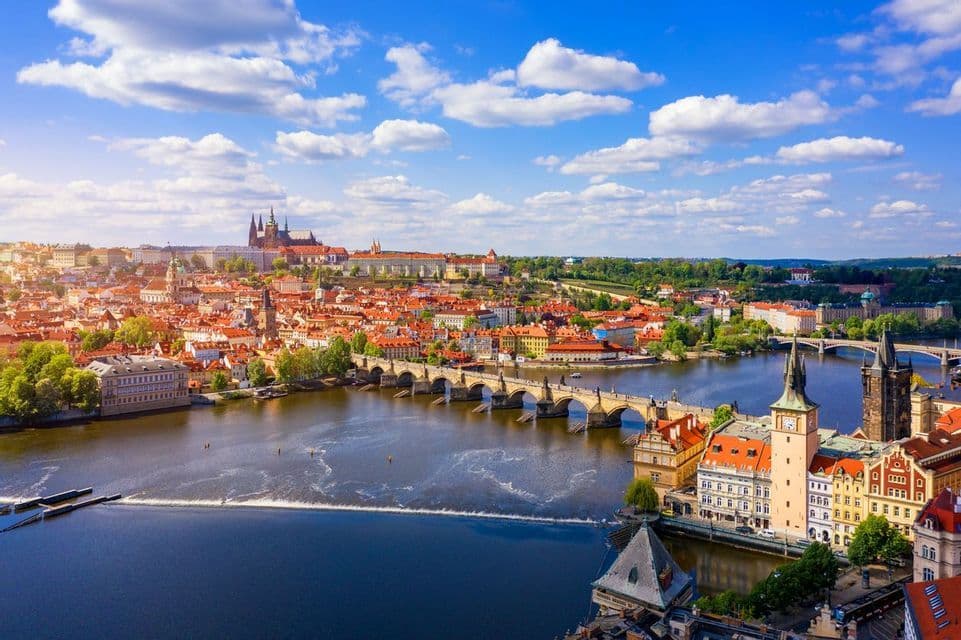 Vue aérienne d'une ville historique avec des toits rouges, un pont en pierre traversant une rivière et un complexe de château sur une colline sous un ciel bleu.