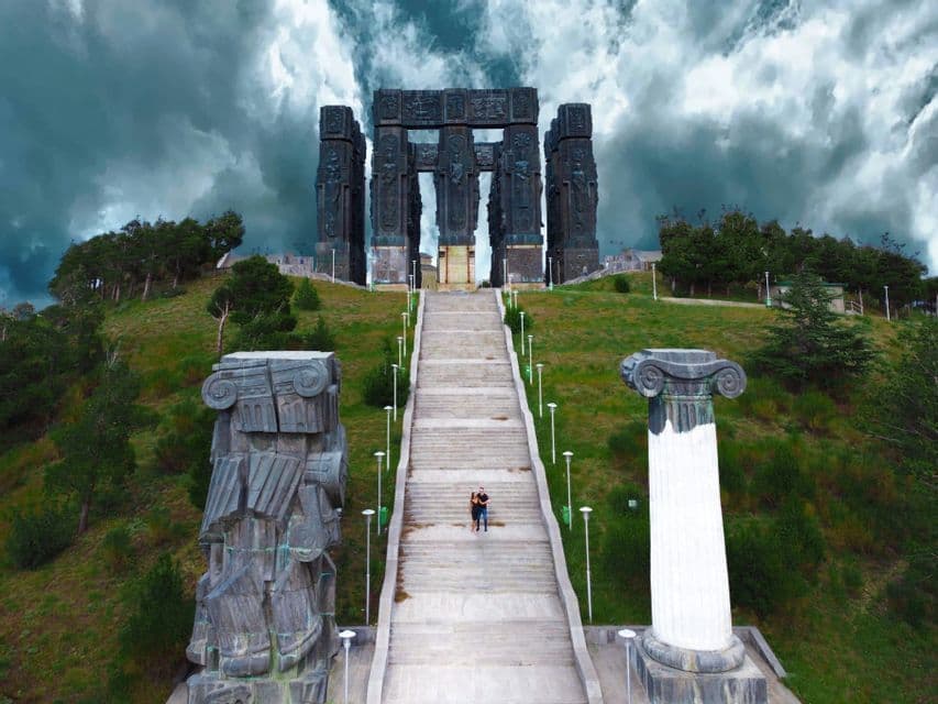 Due persone si trovano su una lunga scalinata in pietra che porta a un imponente monumento su una collina erbosa sotto un cielo tempestoso e nuvoloso.